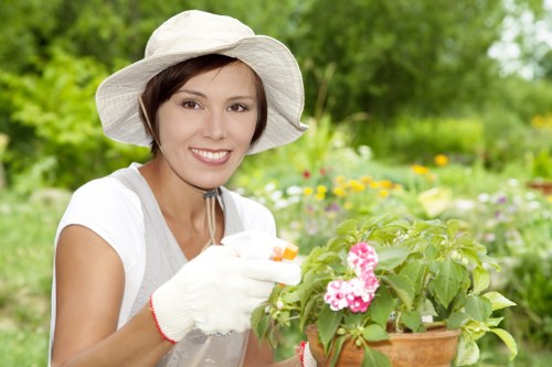 A woman with short brown hair and light skin, wearing a wide-brimmed white gardening hat and white gardening gloves with a red trim, is smiling while tending to a potted flowering plant with pink blooms. She is standing outdoors in a garden with lush green foliage, including various plants and flowers, and a backdrop of trees and shrubs. The scene suggests a pleasant, mild weather day suitable for gardening activities, emphasizing care and maintenance of ornamental plants in a landscaped outdoor space that could be typical of gardens serviced by Gardening Services Dalston in London, combining natural beauty and gardening expertise. The garden features a mix of vibrant and natural tones through the green leaves and colorful flowers, with a well-maintained environment visible in the background.